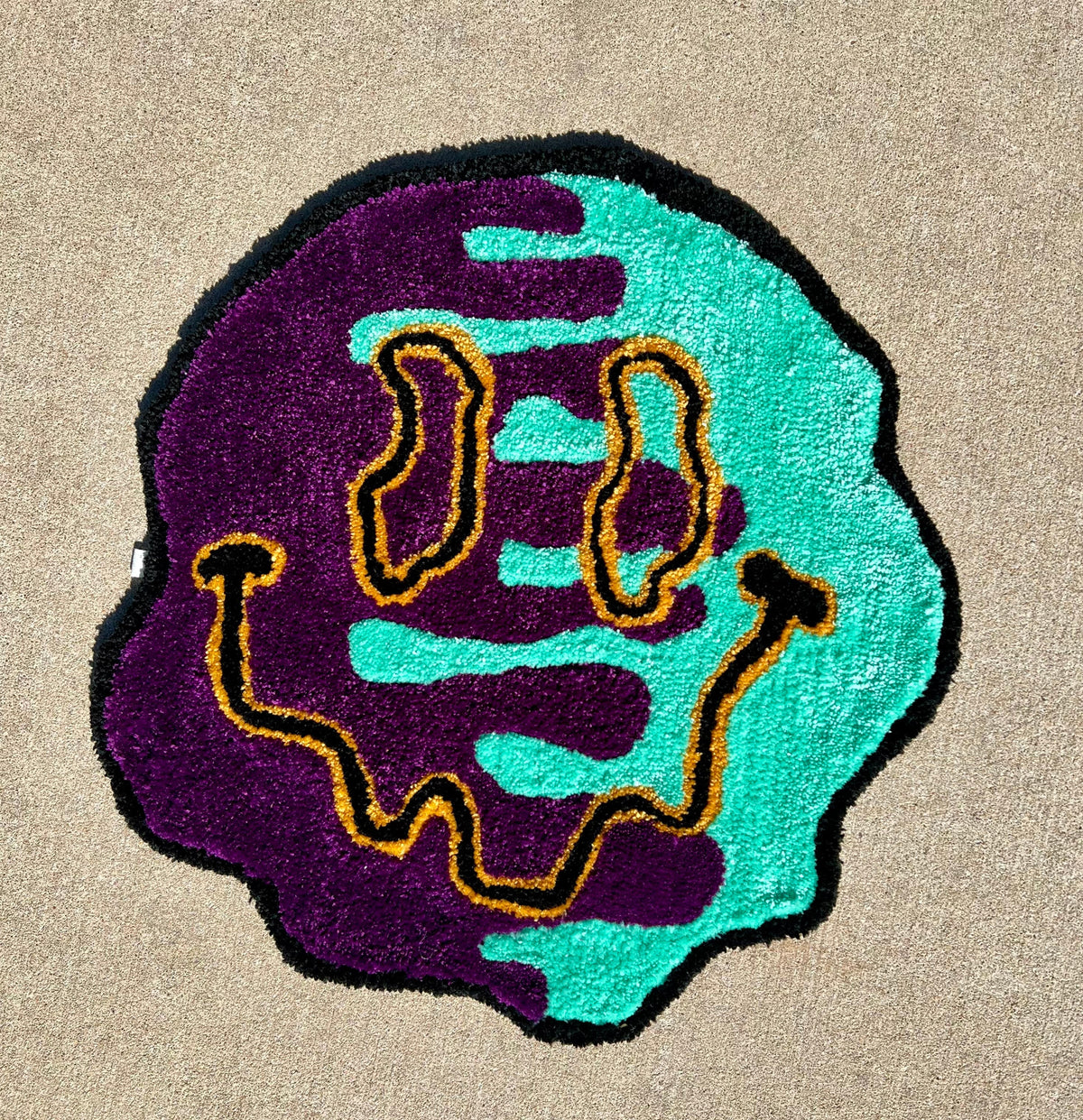 Smiley face rug in black, dark yellow, teal, and dark purple, size 25x25 inches.